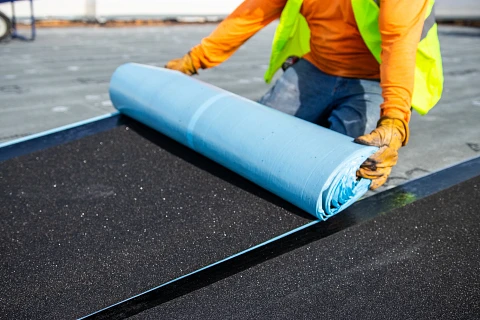 Worker installing commercial roofing material, rolling out blue roofing membrane on a flat surface, demonstrating expert craftsmanship and quality installation standards for THP Roofing of Knoxville.